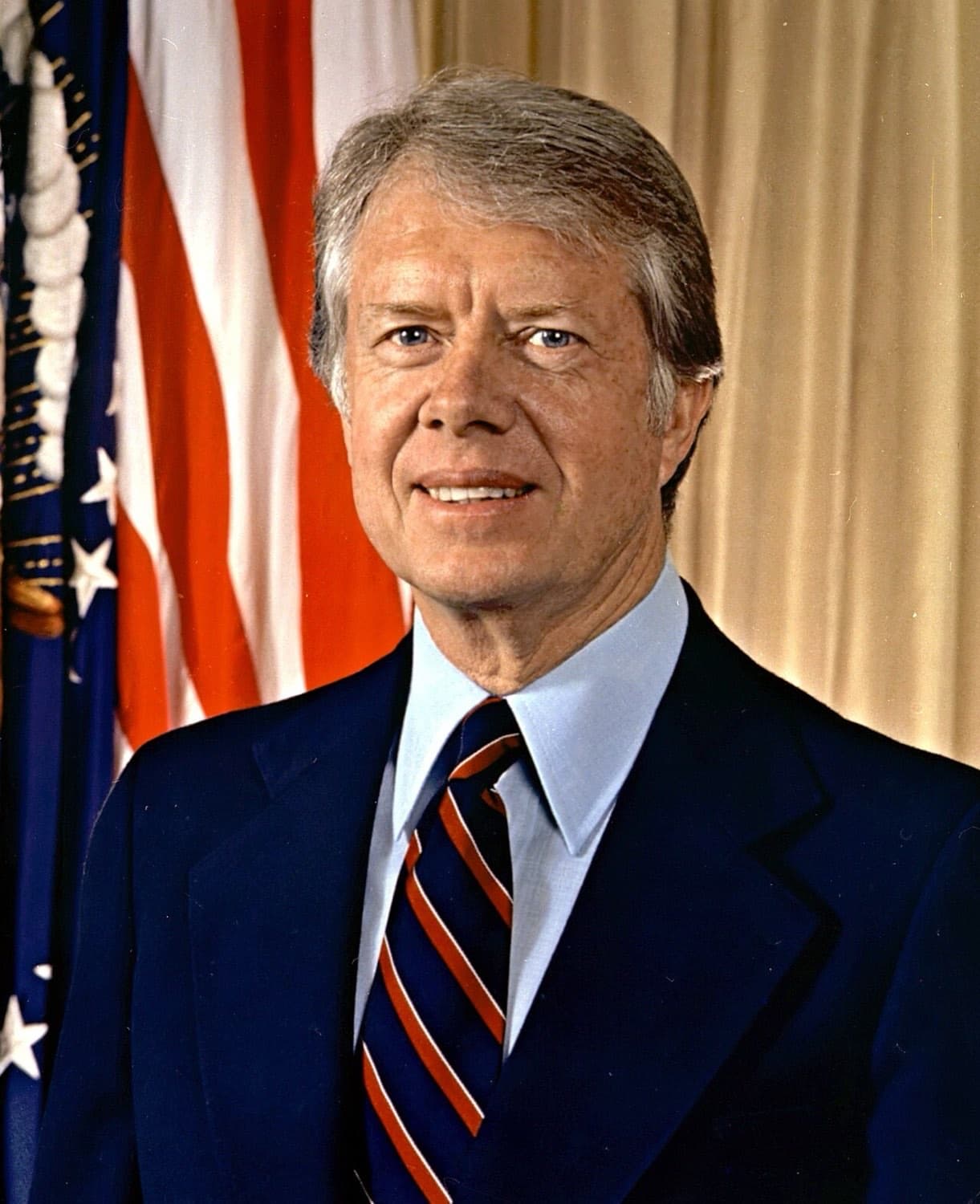 Jimmy Carter in a dark suit and striped tie in front of an American flag