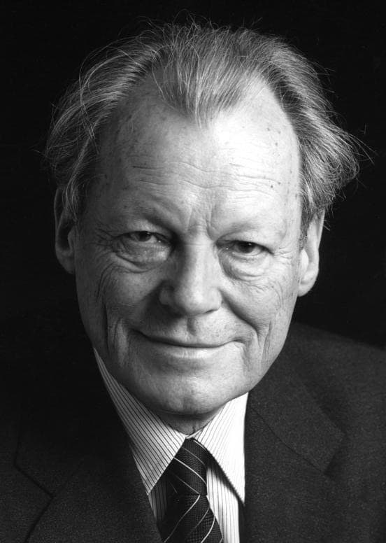 Black and white portrait of Willy Brandt in suit and tie, smiling against a dark background